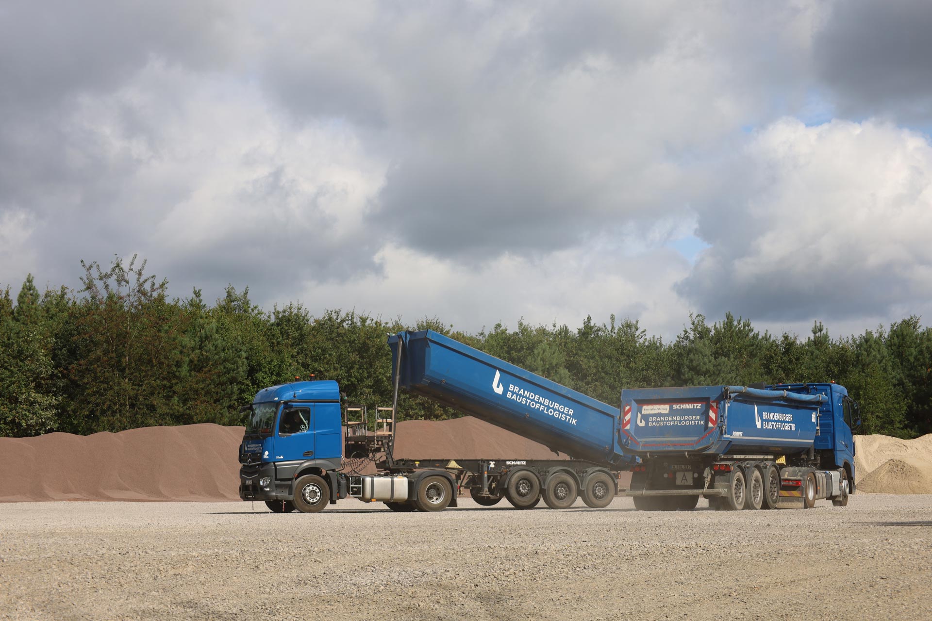 Ein blauer LKW kippt Kies auf einem offenen Lagerplatz bei bewölktem Himmel ab